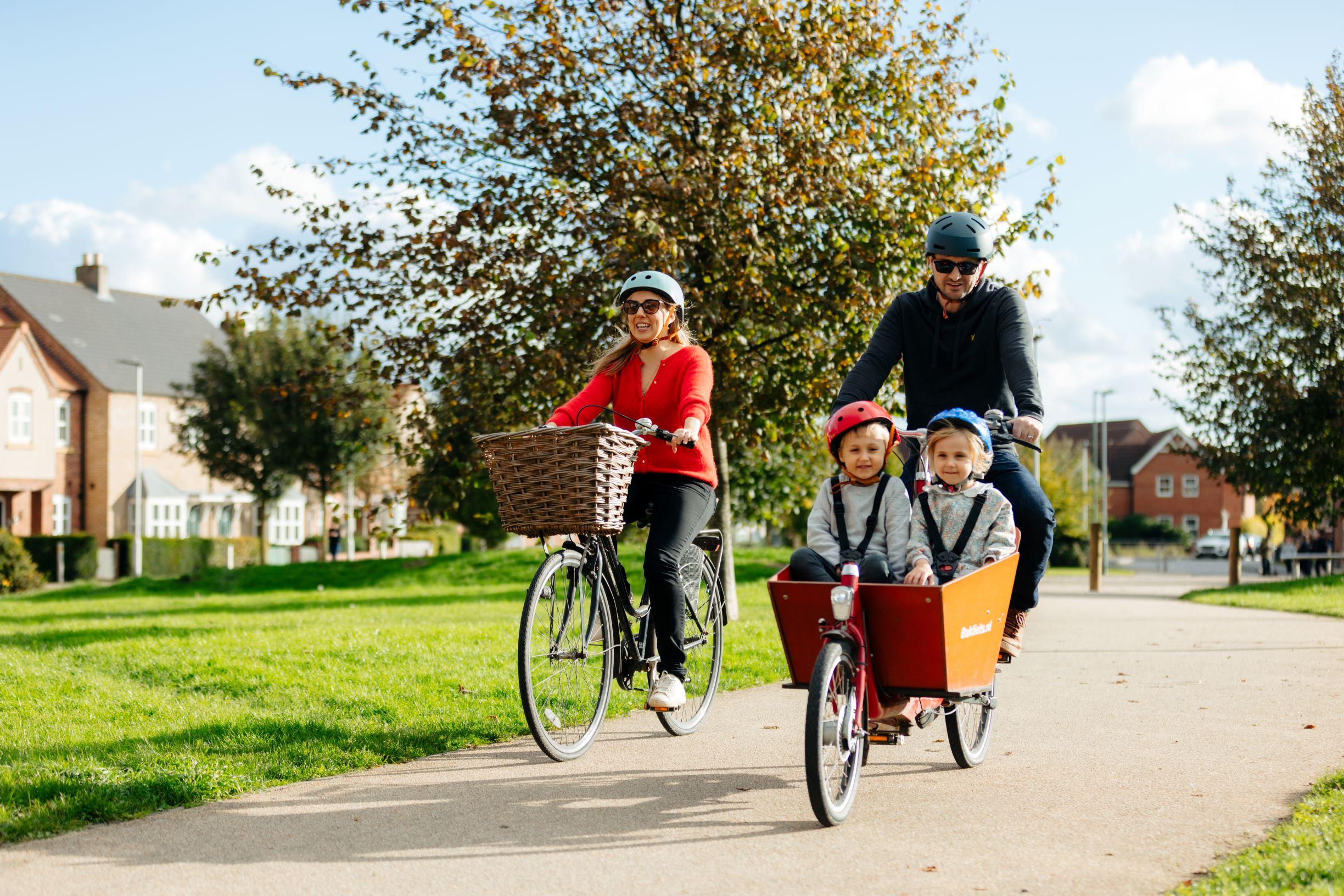 Photograph of a family with 2 young children riding bikes together along a pavement.